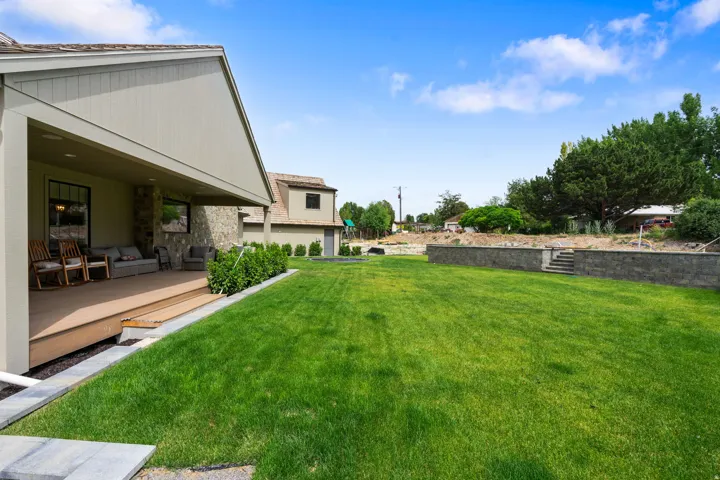 Expansive lawn area featuring a stone retaining wall with integrated steps, a covered patio with a wood-finish floor, and stone accent siding on the dwelling