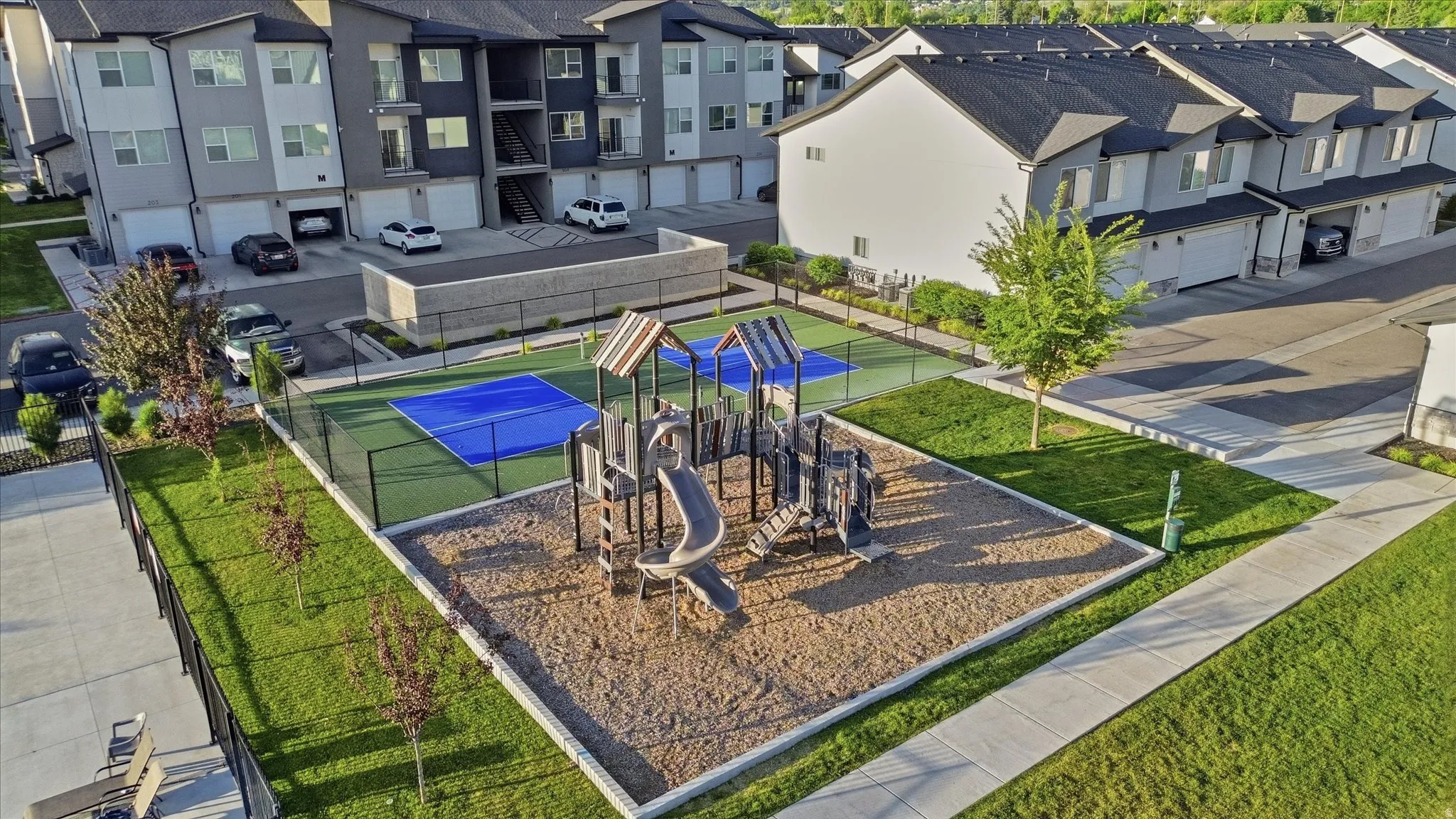 Recreational area featuring a sand-based playground structure with slides and swings, alongside a fenced pickleball court