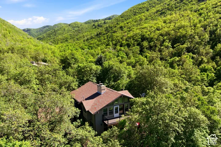 Wooded hillside property with a multi-level structure featuring a brown shingle roof, chimney, and an upper-level balcony