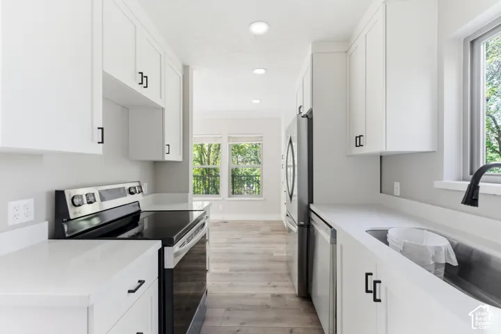 Contemporary kitchen featuring white shaker cabinetry with matte black hardware, stainless steel appliances including a range and refrigerator, light-colored countertops, and wood-finish flooring