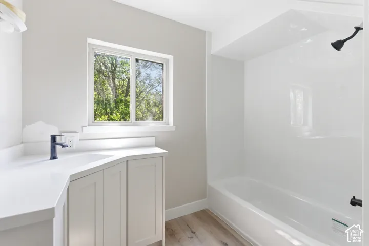 Bathroom featuring a white vanity with integrated sink, a dark-finish single-handle faucet, a window with natural scenery, a white shower/tub combination, and light wood-finish flooring