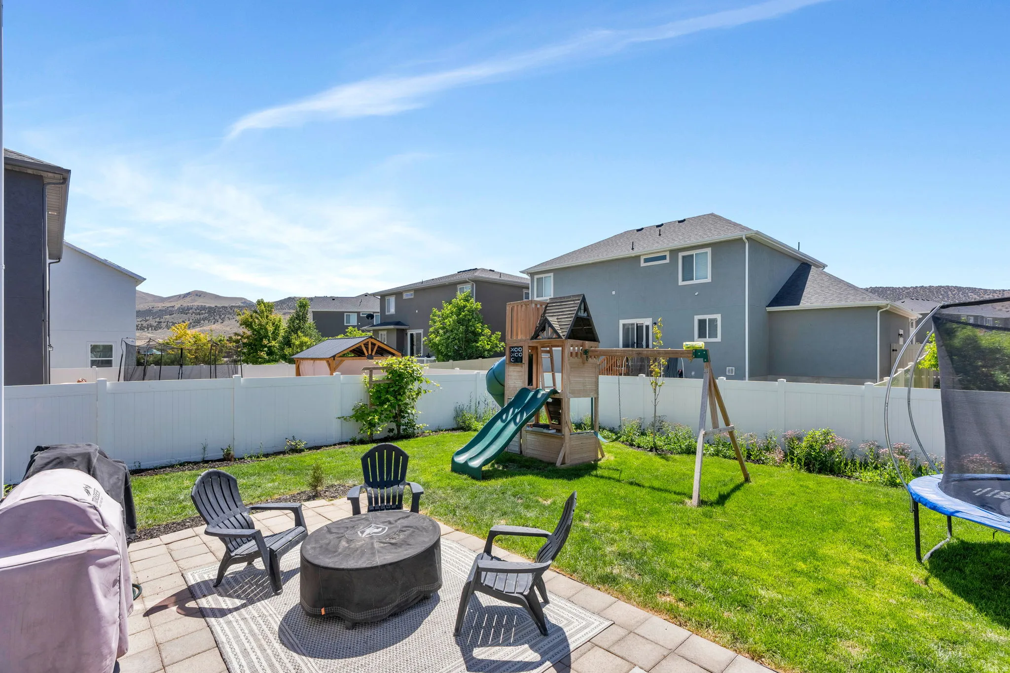 Spacious backyard featuring a paver patio with a fire pit area, manicured lawn, and white vinyl fencing