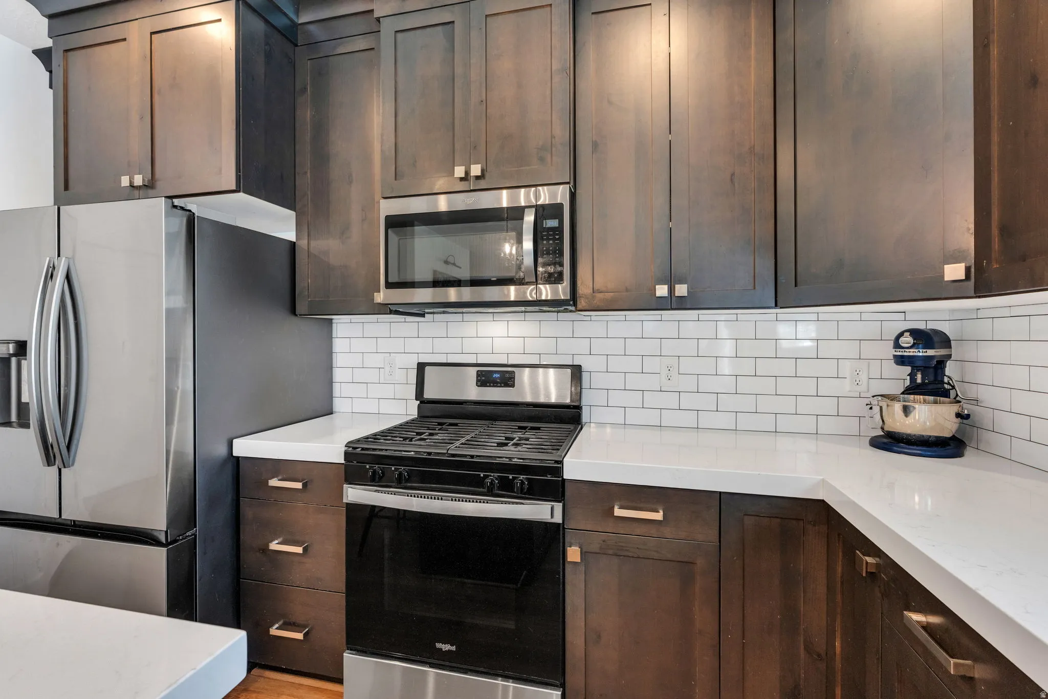 Kitchen featuring dark wood cabinetry, white subway tile backsplash, light-toned countertops, stainless steel appliances, and brass-tone hardware