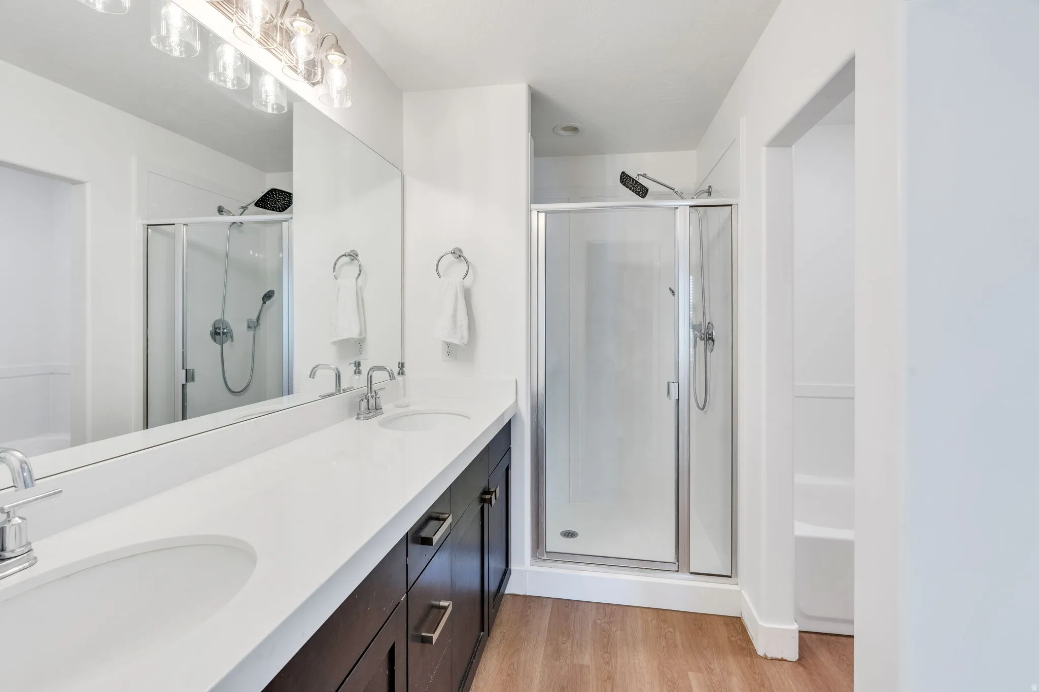 Dual vanity with undermount sinks and chrome fixtures, extensive countertop space, dark wood-finish cabinetry, full-width mirror, and a glass-enclosed shower with a rainfall showerhead