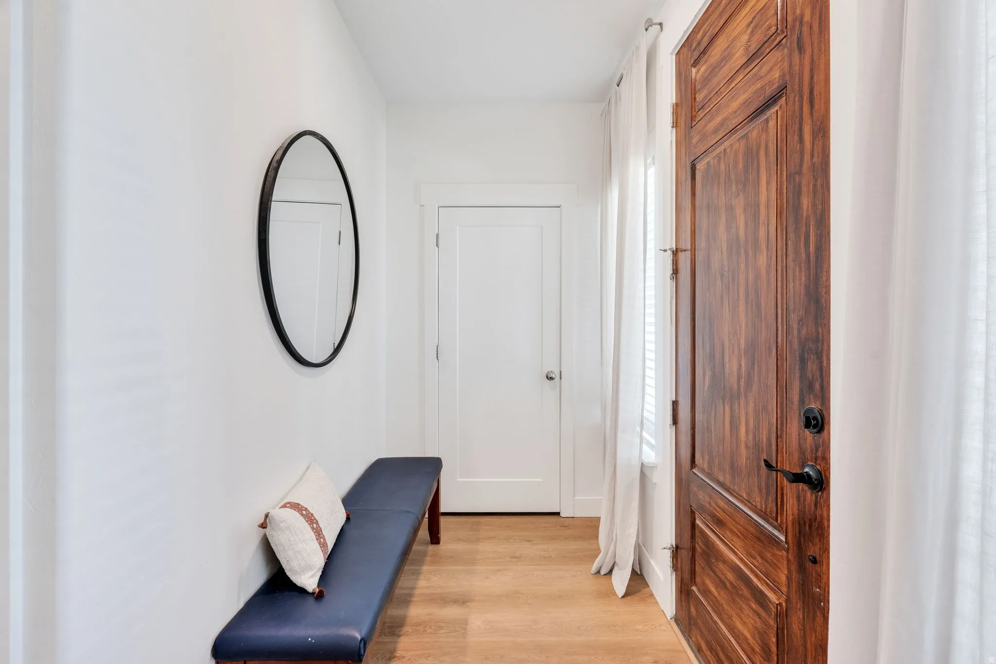 Entryway featuring a rich wood-finish door with black hardware, light wood-finish flooring, white walls, and a round wall mirror