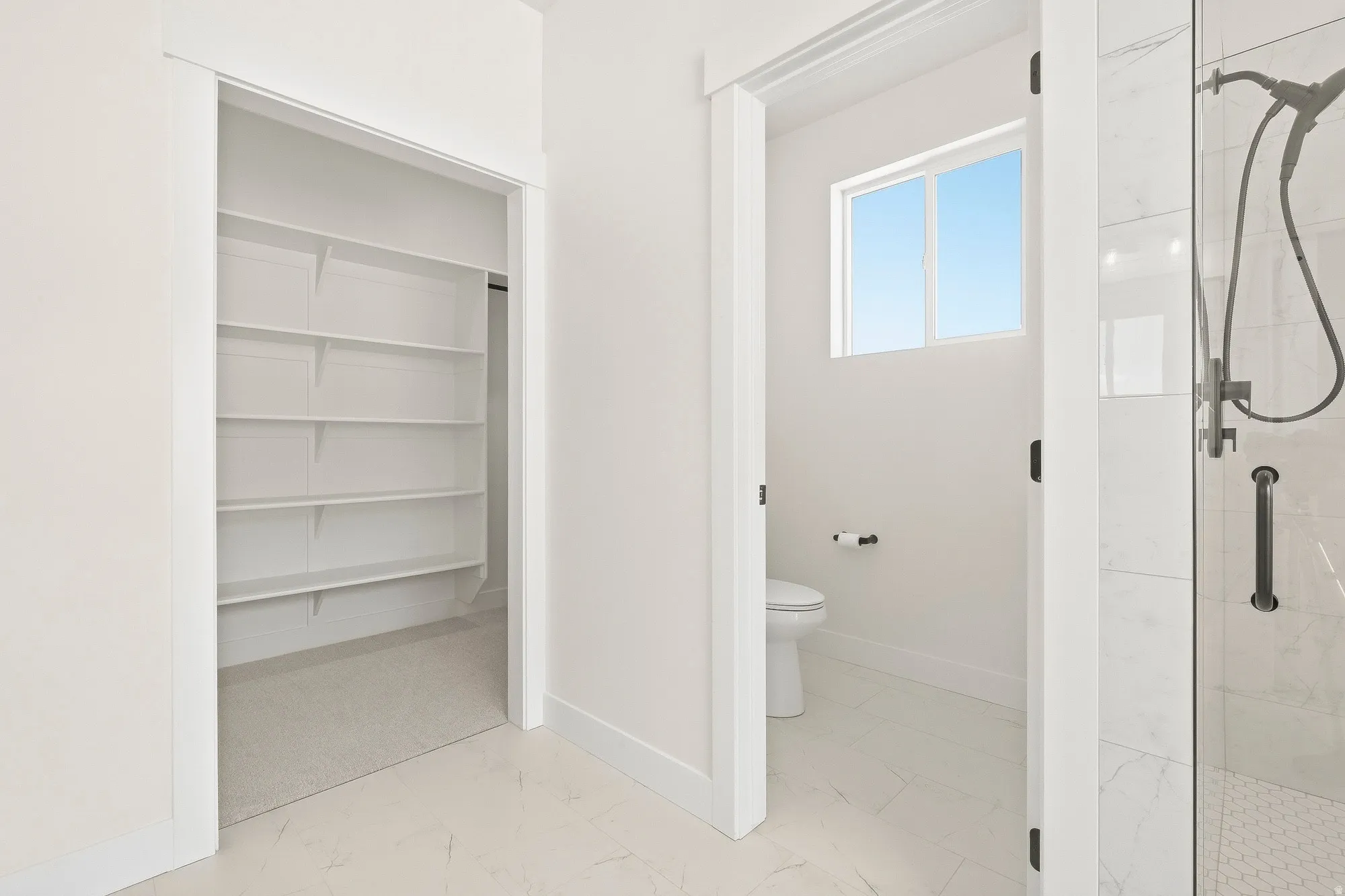 White interior with built-in shelving, a toilet, a window, and a glass-enclosed shower featuring a handheld showerhead