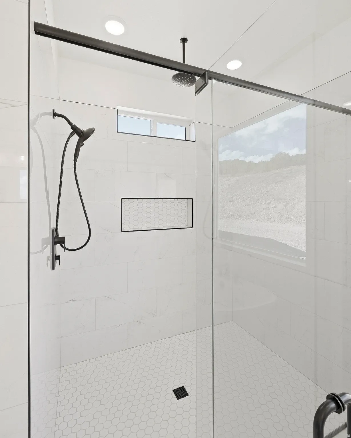 Glass-enclosed shower with a rain shower head, handheld shower, recessed niche with hexagonal tile, and hexagonal floor tiling