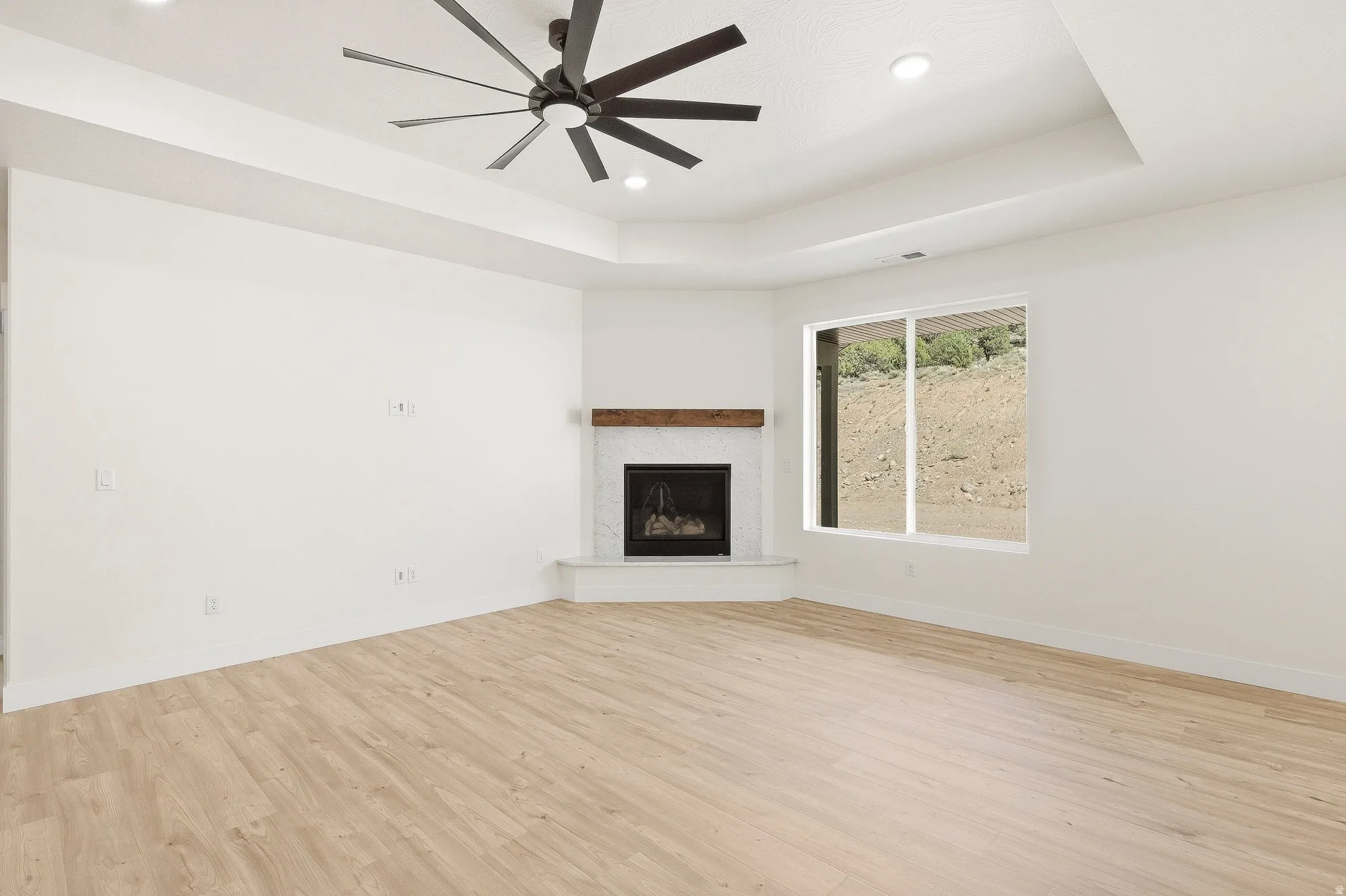 Tray ceiling with recessed lighting and a ceiling fan, a corner fireplace with a white surround and a wood mantel, and wood-finish flooring