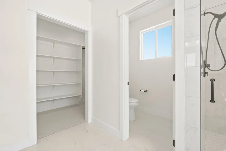 White interior with built-in shelving, a toilet, a window, and a glass-enclosed shower featuring a handheld showerhead