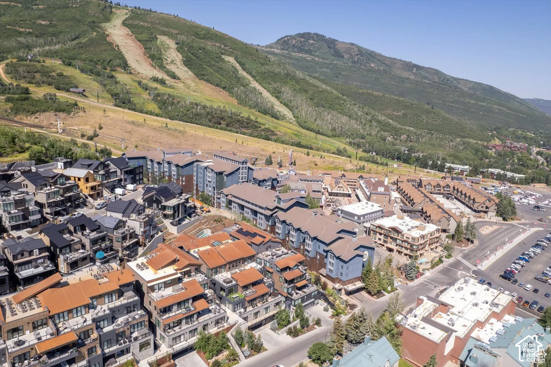 Expansive aerial showcasing a mountain community featuring multi-story residential buildings with diverse architectural styles, a large parking area, and tree-lined slopes