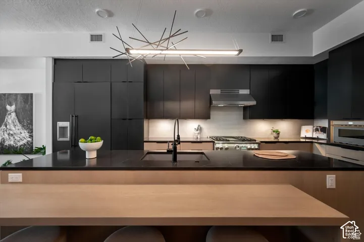 Modern kitchen featuring black flat-panel cabinetry, a wood-finish island with integrated seating, dark countertops, a stainless steel range with a coordinating hood, and a contemporary linear light fixture
