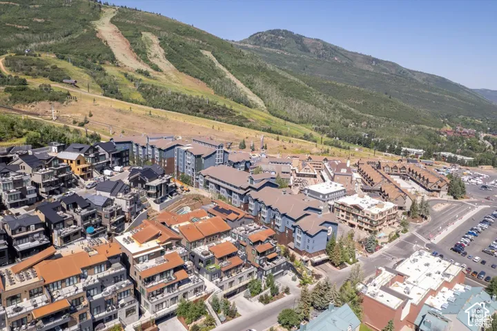 Expansive aerial showcasing a mountain community featuring multi-story residential buildings with diverse architectural styles, a large parking area, and tree-lined slopes