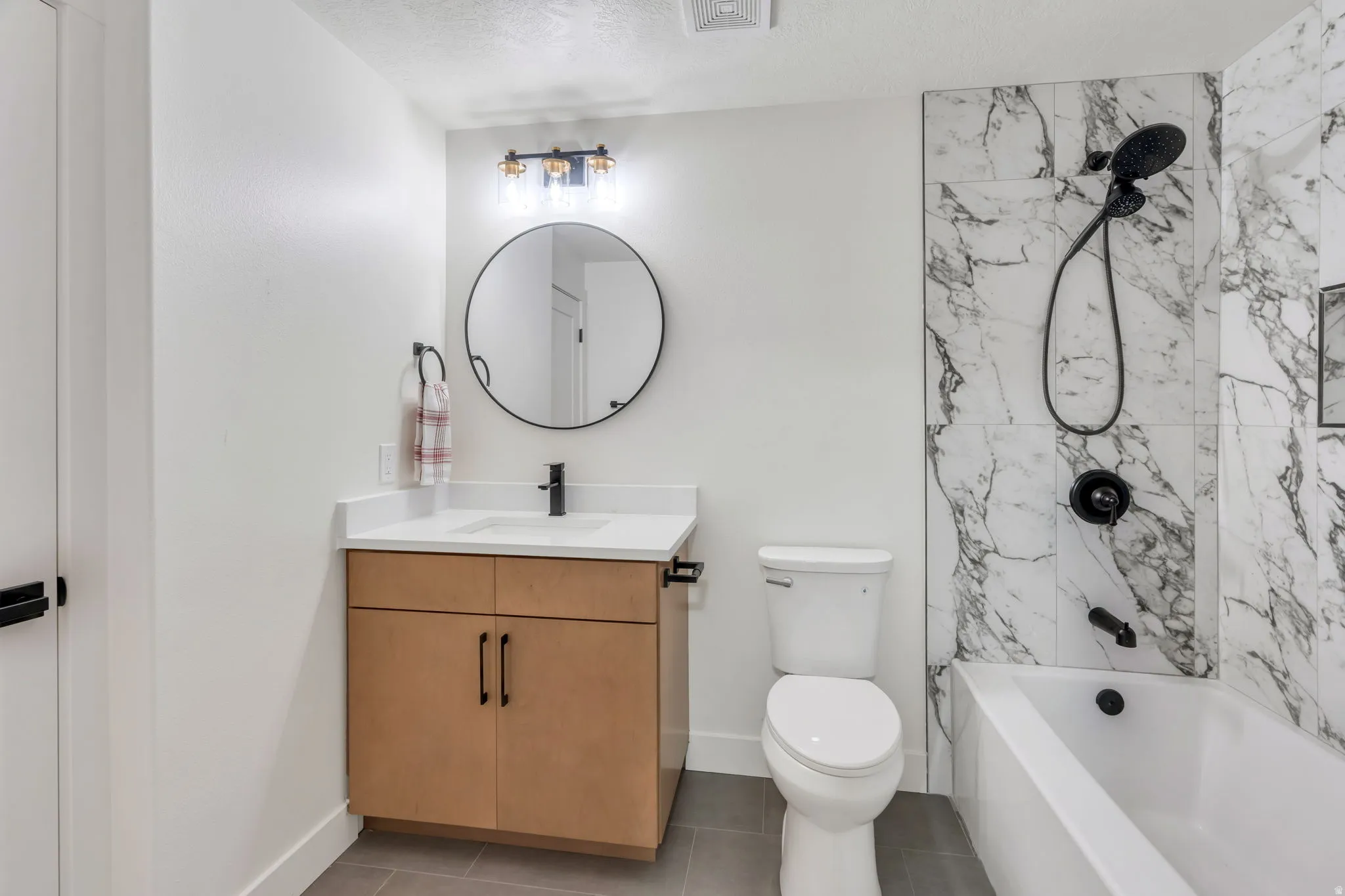 Bathroom featuring a light wood-finish vanity with a white countertop and undermount sink, a circular mirror, and a three-light vanity fixture