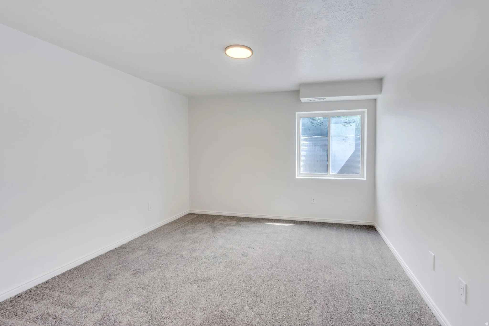 Spacious room featuring light gray carpeting, white walls, a single window, and a wall-mounted climate control unit