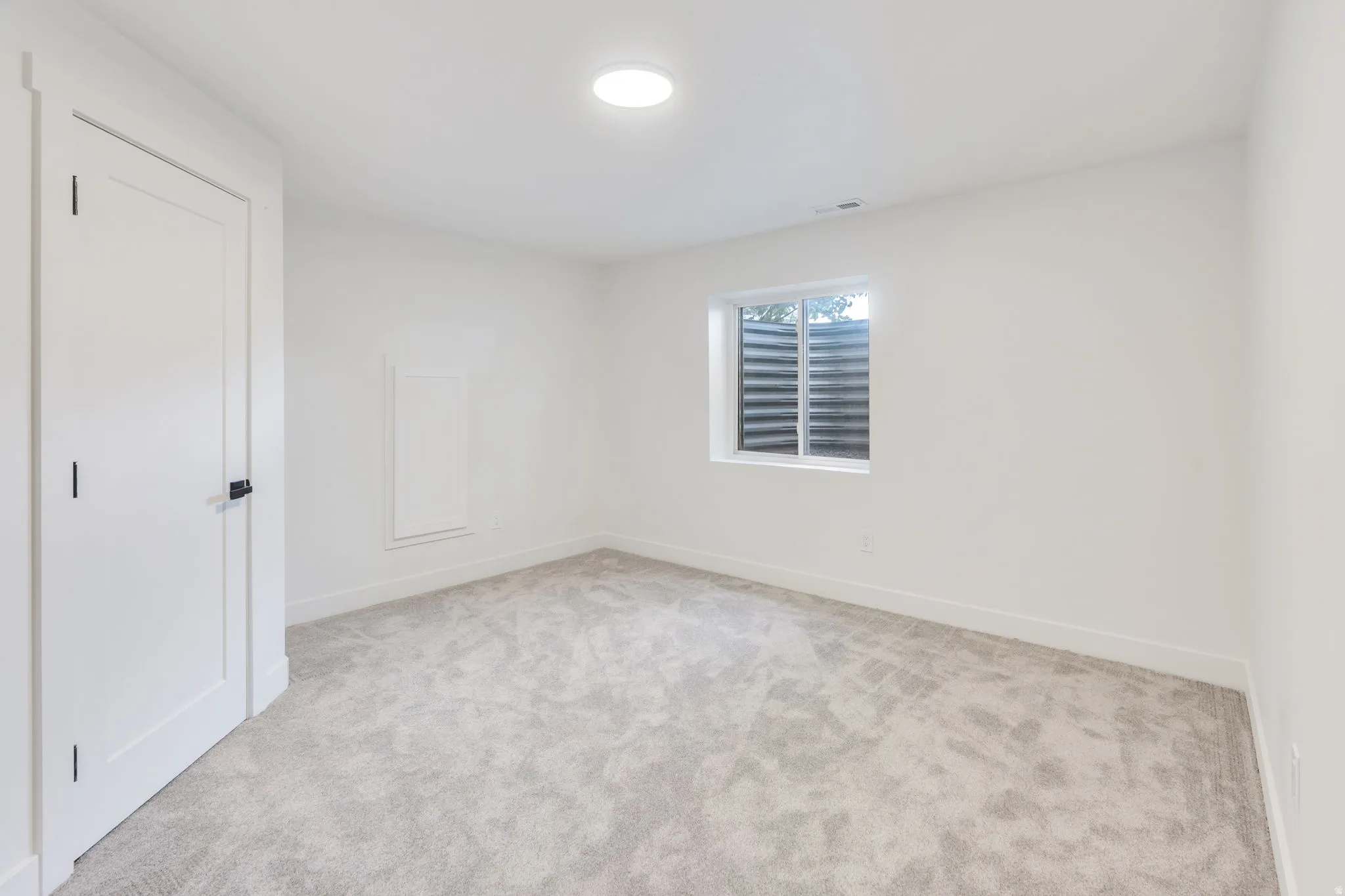 Spacious room featuring light gray carpet, white walls, a white door with dark hardware, a window with horizontal blinds, and a circular recessed ceiling light
