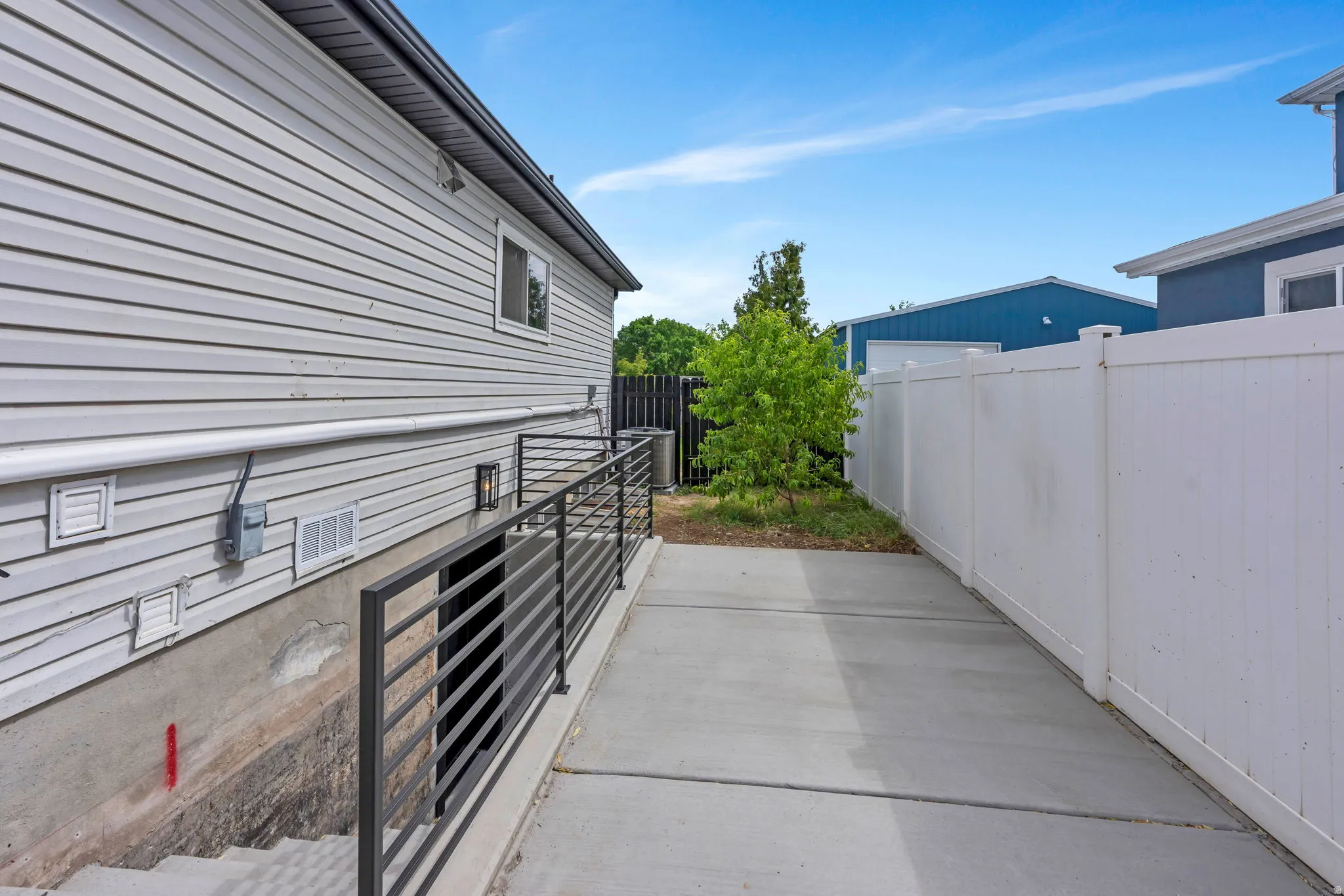 Concrete pathway alongside the home, featuring a white vinyl privacy fence and a dark metal railing leading to a lower-level entrance