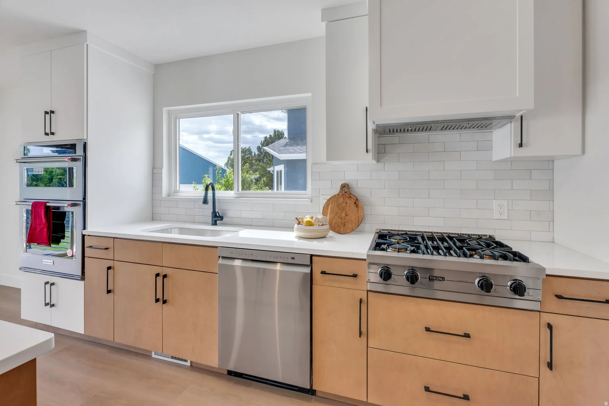 Modern kitchen featuring two-tone cabinetry, stainless steel appliances, a gas range with ventilation hood, white subway tile backsplash, and a large window above the sink
