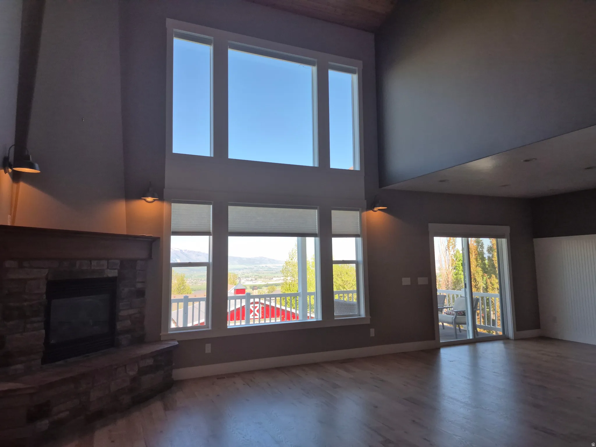 Vaulted ceiling great room featuring expansive window walls, a floor-to-ceiling stone-clad fireplace with wood mantel, and light finish hardwood plank  flooring