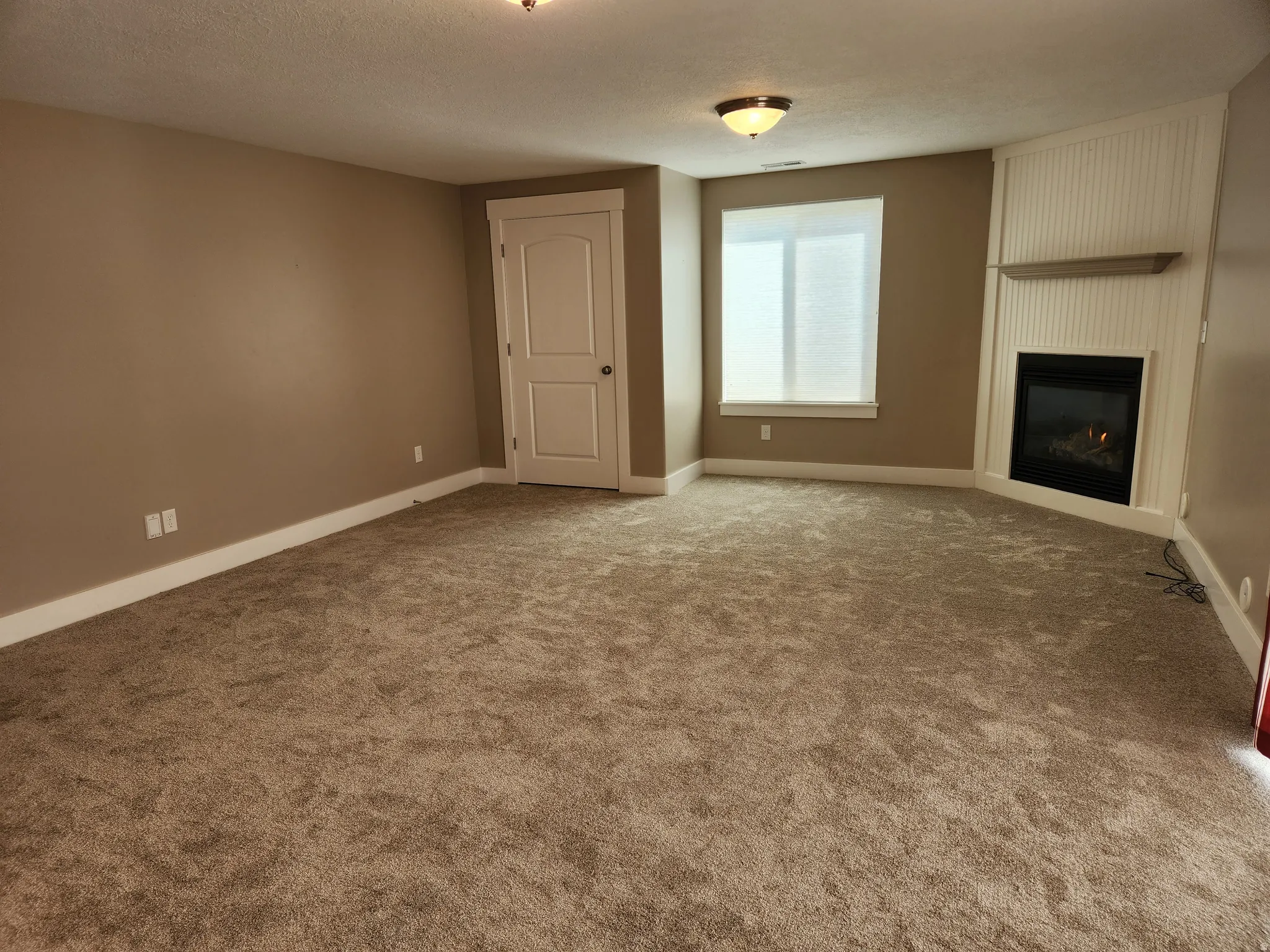 Spacious room featuring plush carpeting, a corner gas fireplace with a white beadboard surround and mantel shelf, a large window, and light tan wall color, open to the 2nd kitchen.  Room has exterior entrance.