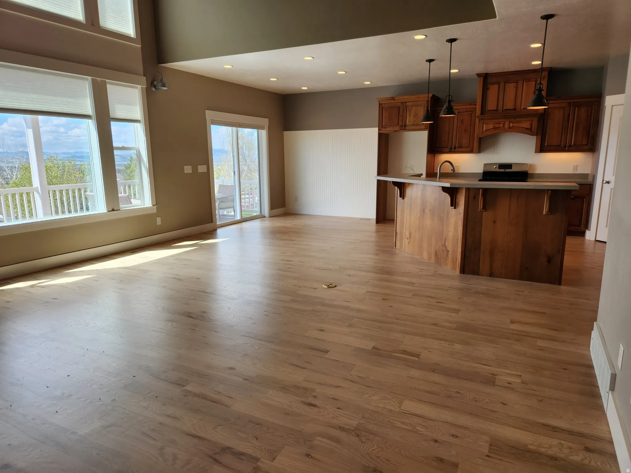 Open-concept living area featuring newly refinished hardwood plank flooring, a kitchen island with built-in cabinetry, stainless steel appliances, and abundant natural light from large windows and a sliding glass door