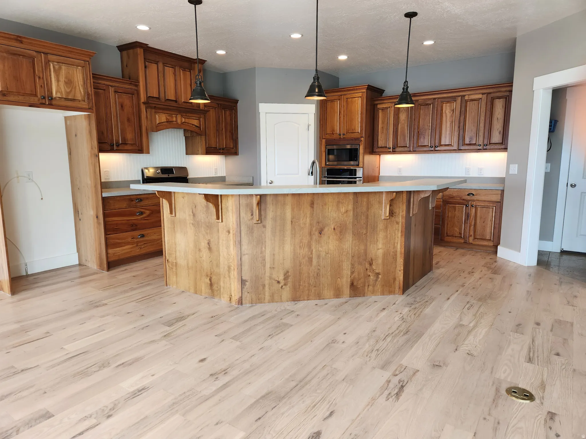Spacious kitchen featuring an expansive island, light finish hardwood flooring, custom cherry cabinetry, under-cabinet lighting, and integrated stainless steel appliances