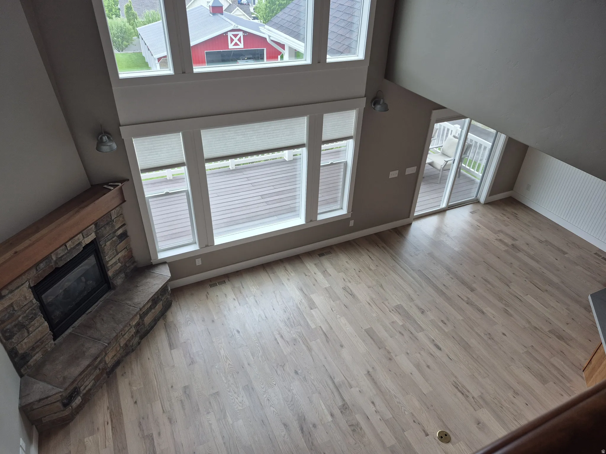 Open-concept living area featuring hardwood plank flooring, a stone-surround fireplace with a wood mantle, high ceilings, multiple windows, and sliding glass door access to an exterior deck