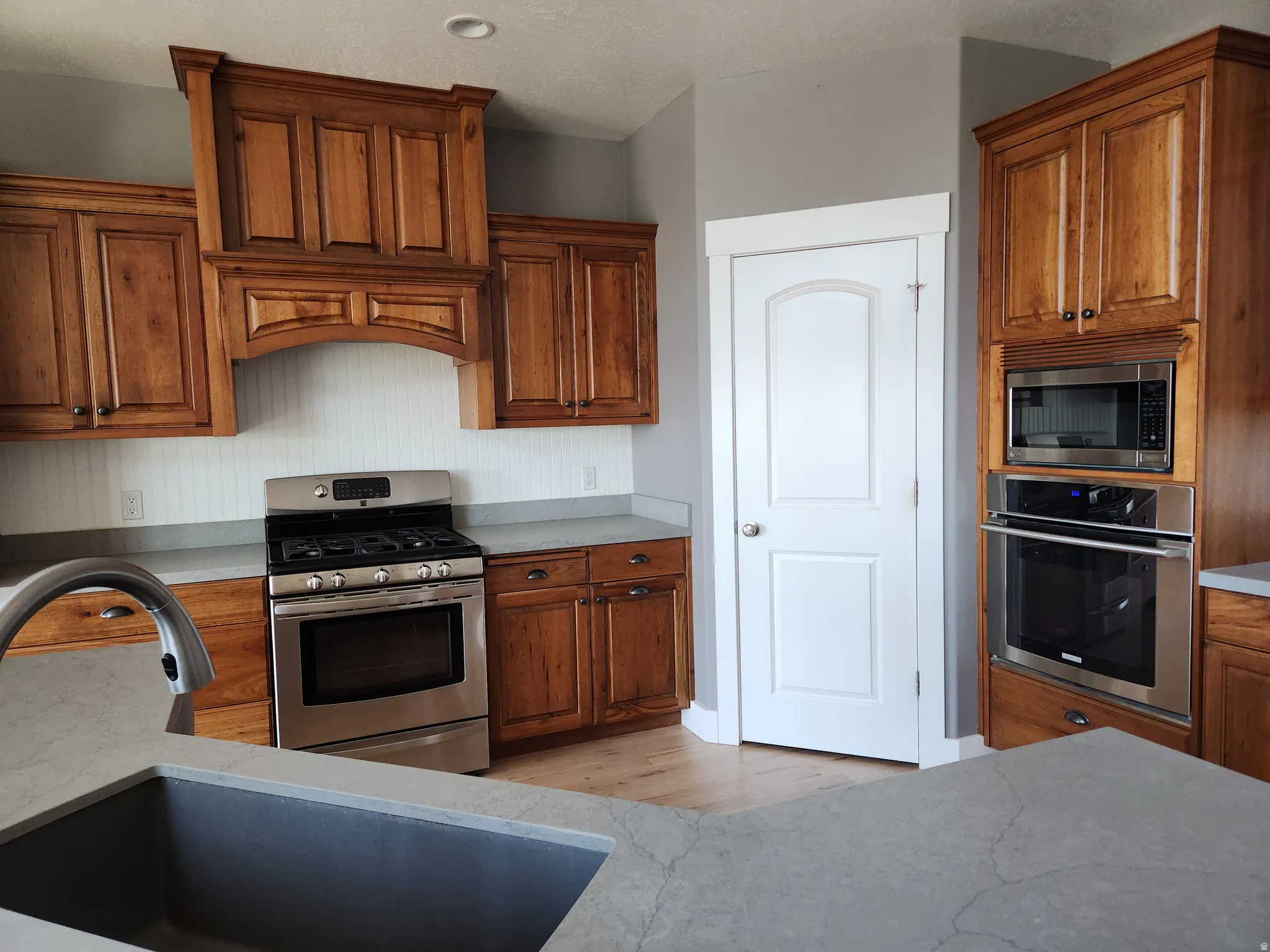 Primary kitchen featuring custom cherry wood cabinetry, stainless steel appliances, a built-in wall oven and microwave, light gray quartz countertops, and a white beadboard backsplash. Walk-in pantry. 2nd kitchen in the walk-out basement level.
