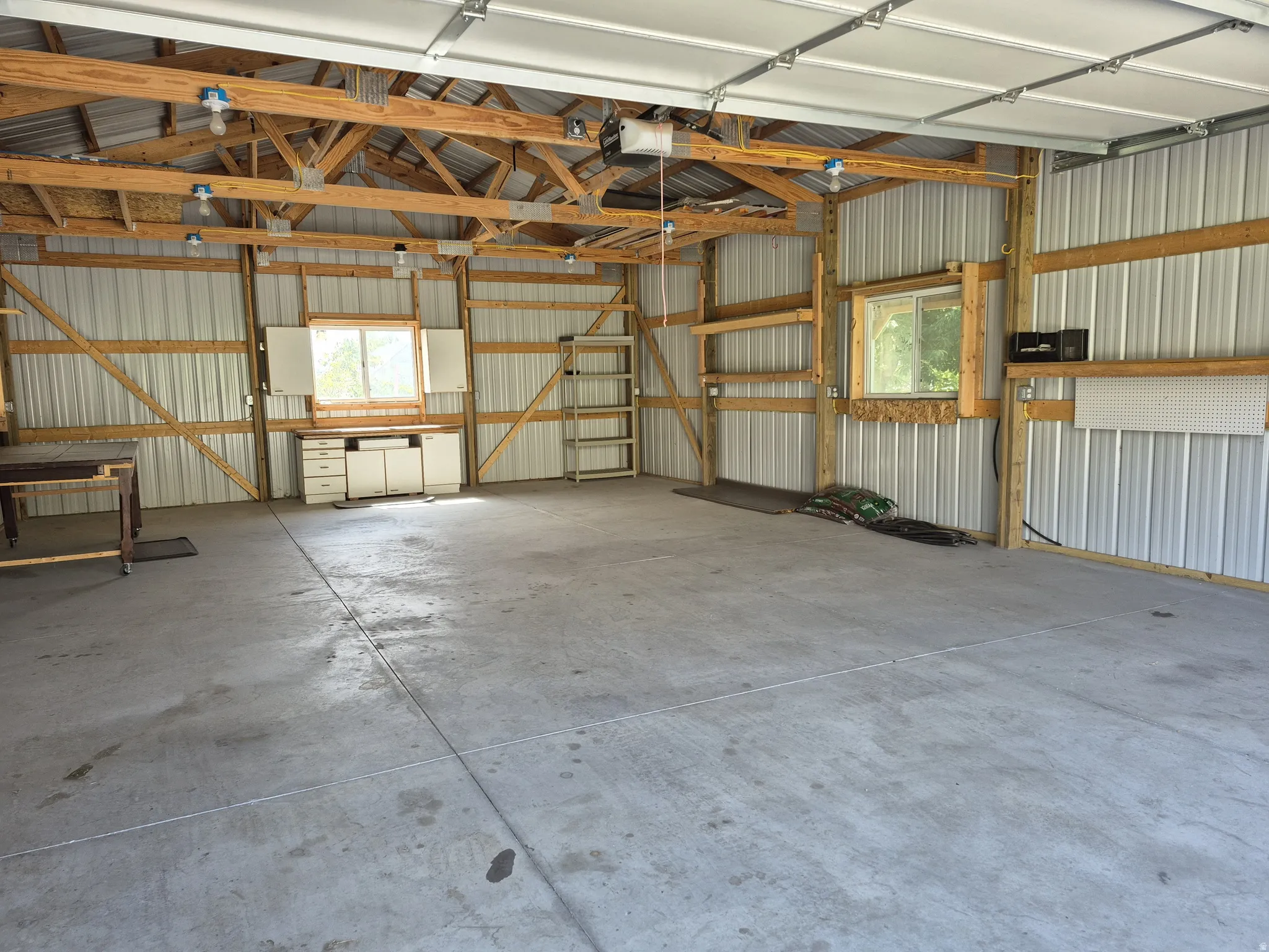 Spacious 26x35' garage interior featuring a concrete floor, exposed wood beam ceiling, electrical outlets and lighting, windows, side man-door, and corrugated metal walls