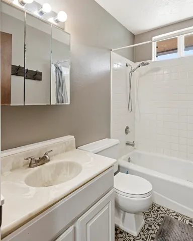 Bathroom featuring a single vanity with an integrated sink and brushed nickel faucet, a medicine cabinet with overhead lighting, a toilet, a bathtub with a shower system and white tiled surround, and patterned floor tiling