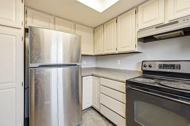 Kitchen featuring light-toned cabinetry, light-colored countertops, a stainless steel refrigerator, a black electric range, and a range hood