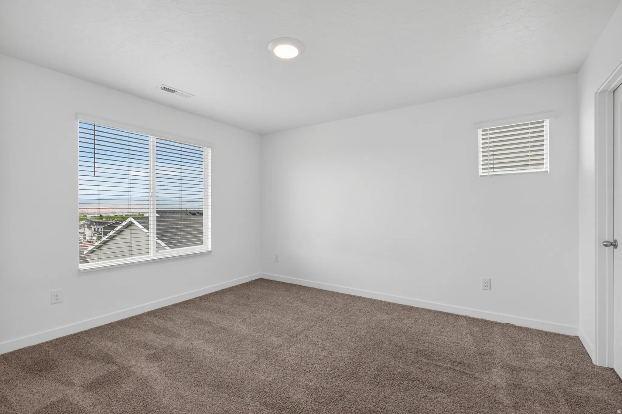 Room with neutral-toned carpeting, white walls, a large window with horizontal blinds, and an additional smaller window