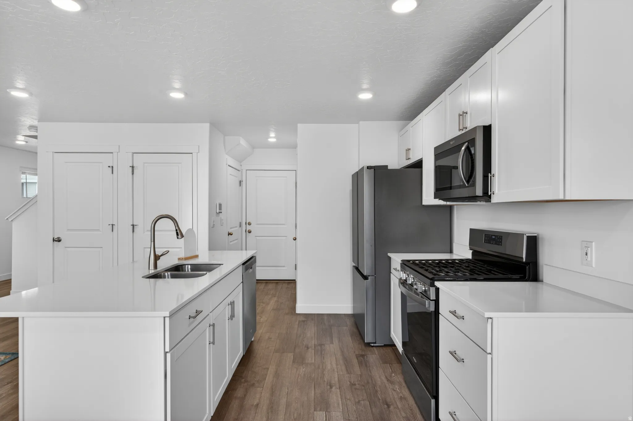 Modern kitchen featuring an island with an undermount double sink, white cabinetry with brushed nickel hardware, stainless steel appliances, white countertops, and wood-finish flooring