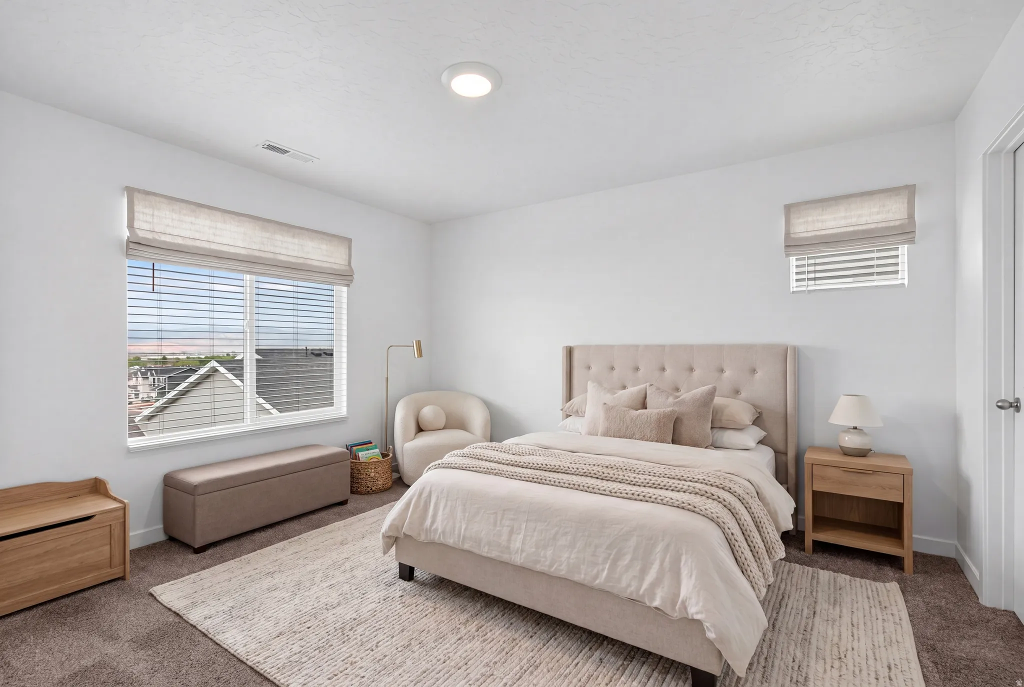 Neutral-toned bedroom featuring crisp white walls, a large window with blinds and a roman shade, an overhead flush-mount light, and dark carpet flooring