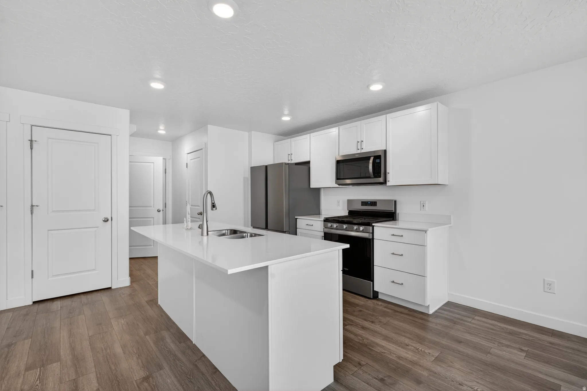 Kitchen featuring a central island with an integrated sink, white shaker cabinetry, stainless steel appliances, wood-finish flooring, and recessed lighting