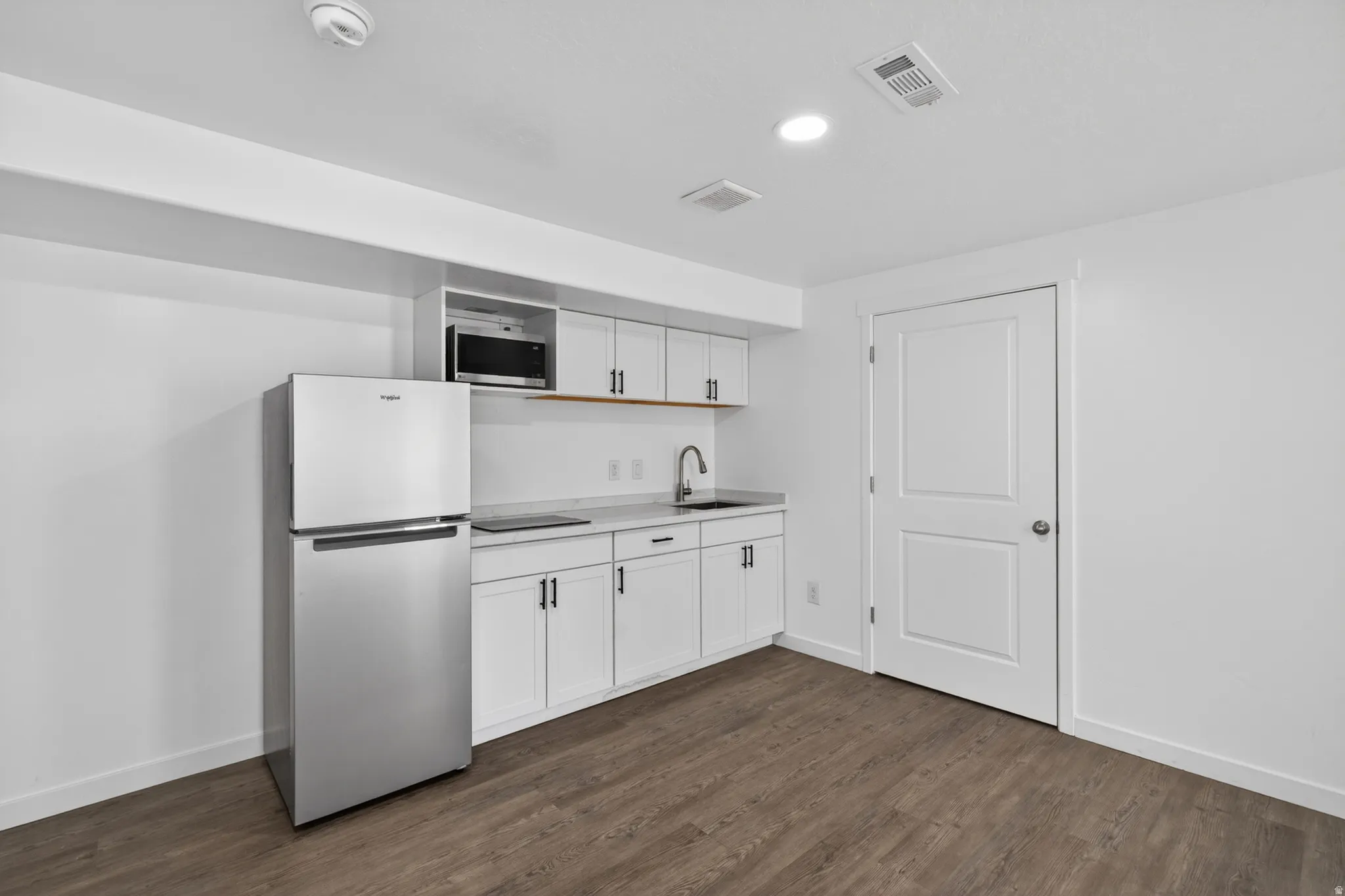 Compact kitchen area featuring a stainless steel refrigerator, white cabinetry with dark hardware, a stainless steel sink with a gooseneck faucet, a built-in microwave, and wood-finish flooring