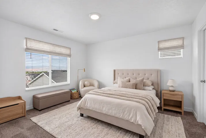 Neutral-toned bedroom featuring crisp white walls, a large window with blinds and a roman shade, an overhead flush-mount light, and dark carpet flooring