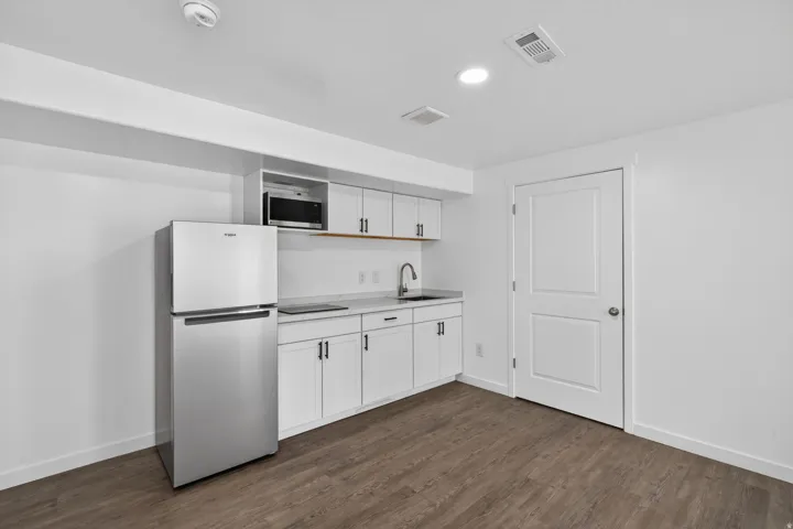 Compact kitchen area featuring a stainless steel refrigerator, white cabinetry with dark hardware, a stainless steel sink with a gooseneck faucet, a built-in microwave, and wood-finish flooring