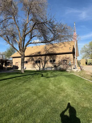 Expansive green lawn with mature tree, brick exterior structure, brown shingle roof, multiple windows, and a flagpole