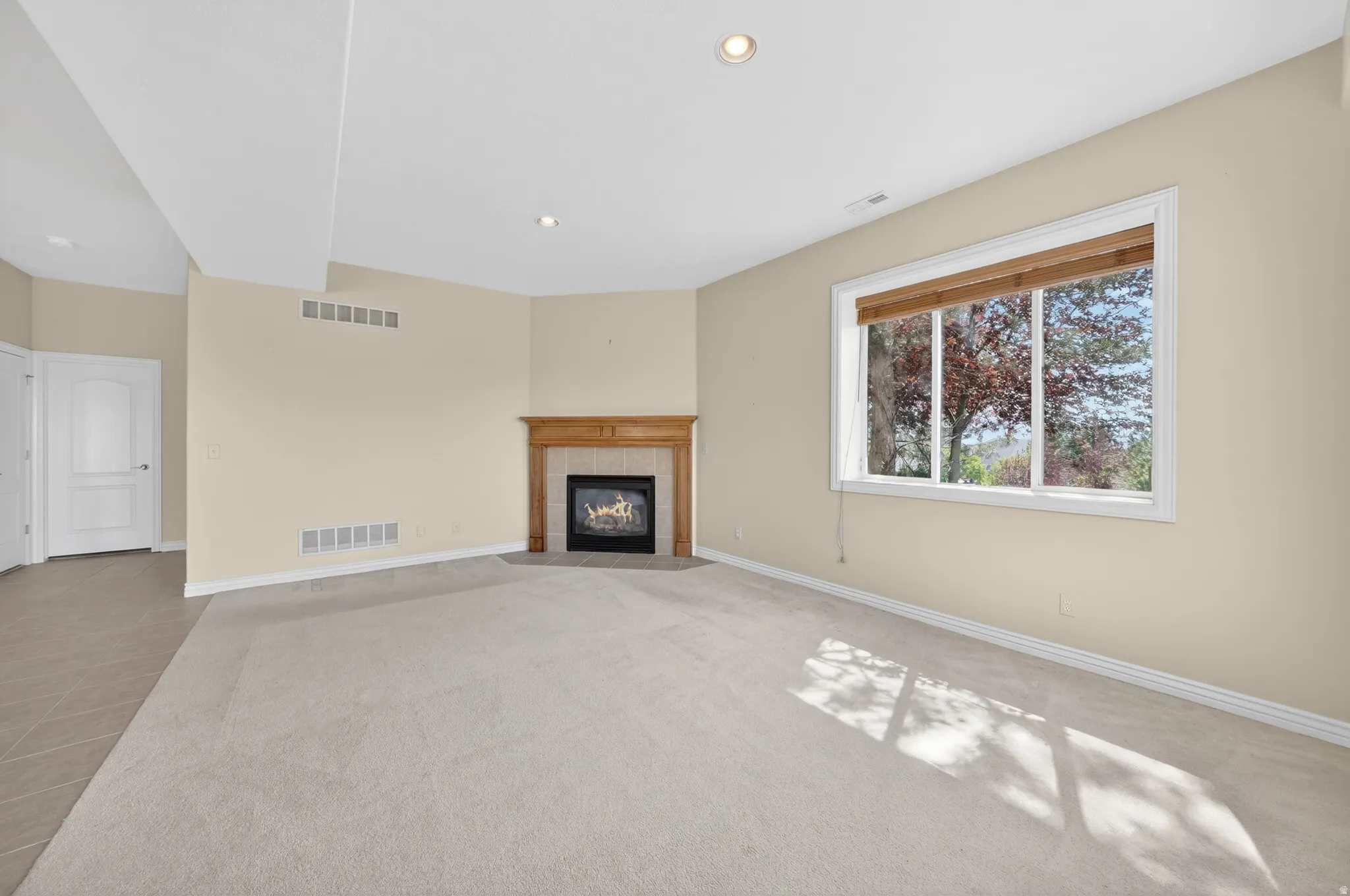 Spacious room featuring neutral carpeting, a corner fireplace with a light wood mantel, a large window with natural light, recessed lighting, and a neutral paint palette