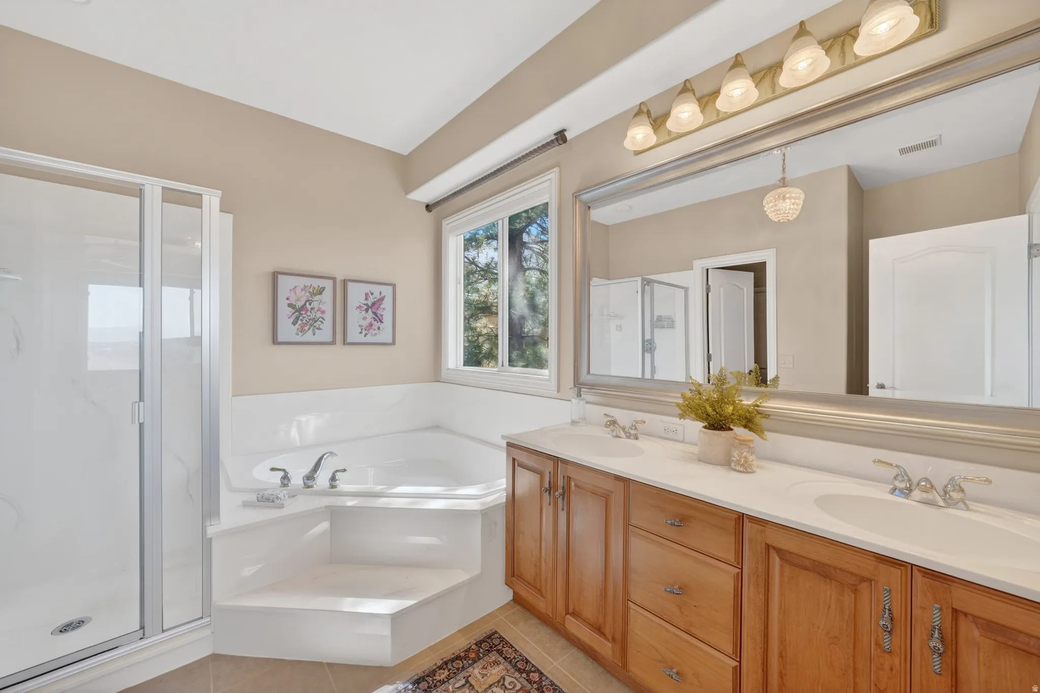 Spacious primary bathroom featuring a corner soaking tub, separate glass-enclosed shower, dual vanity with light wood cabinetry, and a large wall-mounted mirror