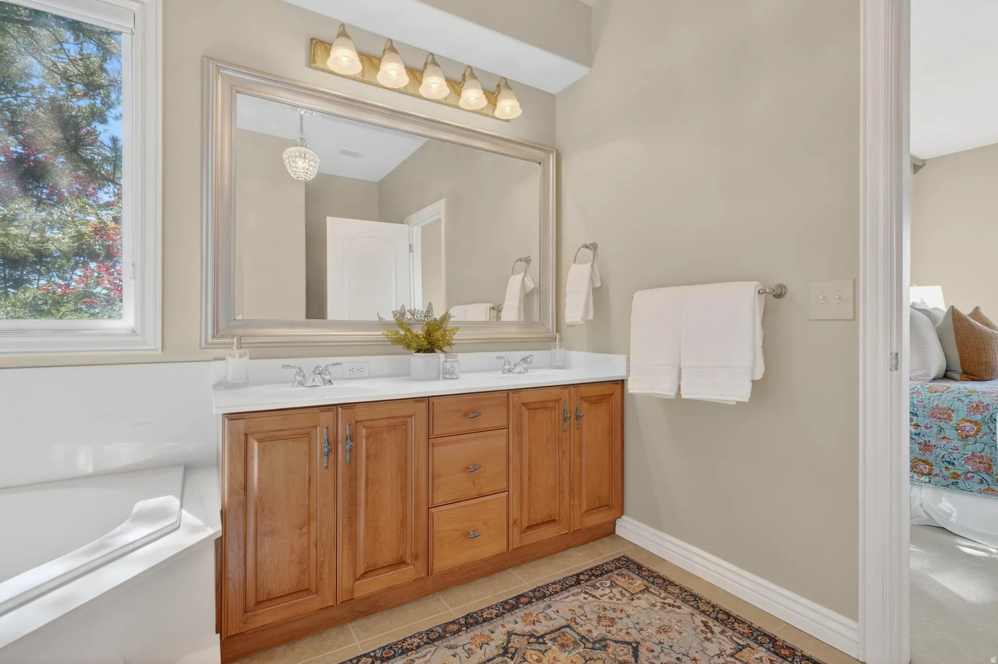 Bathroom featuring a light wood-finish double vanity with white countertops, a large framed mirror, and a built-in soaking tub with an adjacent window