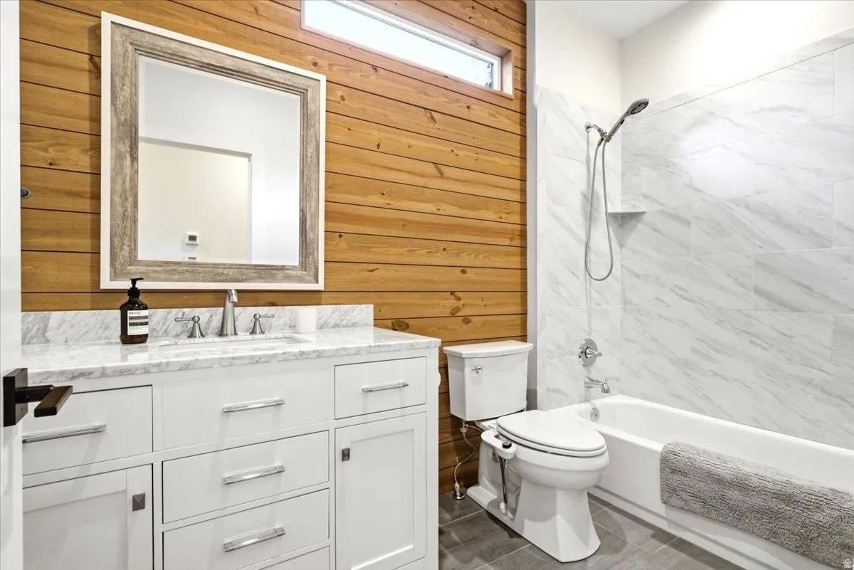 Bathroom featuring a white vanity with a marble countertop and undermount sink, wood-paneled accent wall, a framed mirror, a tub/shower combination with marble-look tile surround, and grey floor tiles