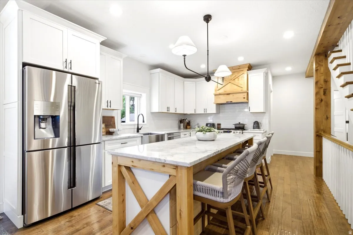 Kitchen featuring a substantial island with a stone countertop and wood-finish base, stainless steel refrigerator, white cabinetry, wood-finish flooring, and a decorative wood range hood
