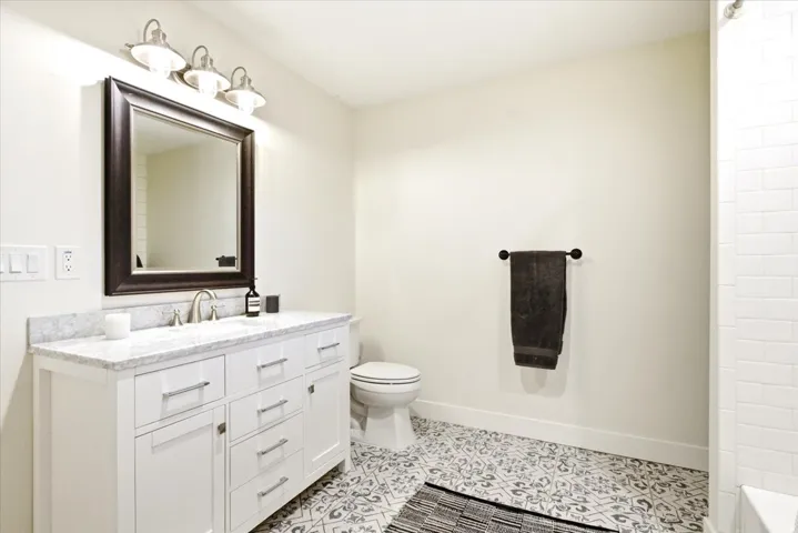Bathroom featuring a white vanity with a stone countertop, a framed mirror, a three-light vanity fixture, patterned floor tiles, and subway tile shower surround