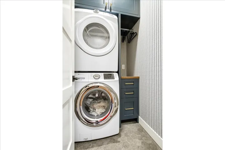 Laundry area featuring a stacked washer and dryer, custom cabinetry with a wood-finish countertop, hexagonal floor tiles, and striped wallpaper