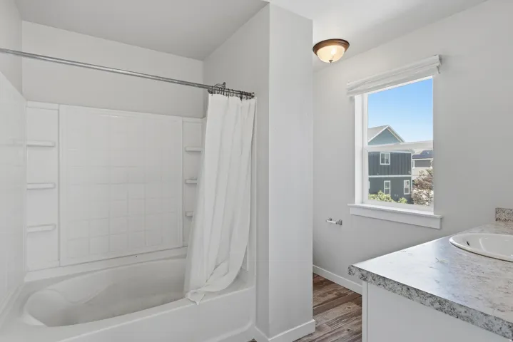 Bathroom featuring a white tub-shower combination with integrated shelving, wood-finish flooring, a window with natural light, a vanity with a drop-in sink, and a speckled countertop