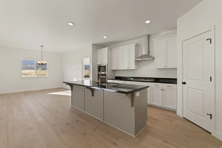 Contemporary kitchen featuring an expansive central island with dark countertops, white cabinetry with gold-tone hardware, stainless steel range hood, and wood-finish flooring