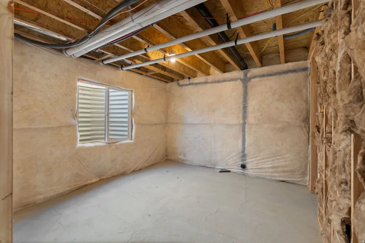 Unfinished room featuring a concrete floor, exposed ceiling joists, insulation-wrapped walls, an egress window, and visible utility lines