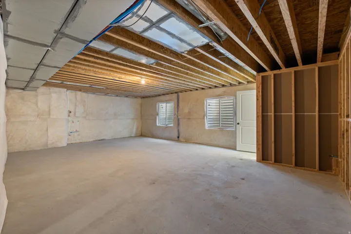 Unfinished basement featuring exposed wood joists, concrete flooring, two windows with louvered coverings, a white two-panel door, and framing for additional rooms