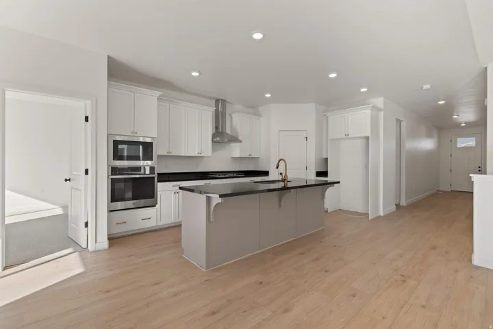 Open-concept kitchen featuring an island with a dark countertop and gold-toned faucet, stainless steel appliances, white cabinetry, and wood-finish flooring